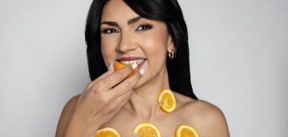 A woman smiles while holding a fresh orange slice and wearing more on her body, promoting natural beauty and nutrition.