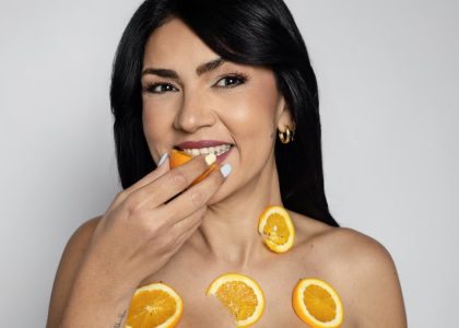 A woman smiles while holding a fresh orange slice and wearing more on her body, promoting natural beauty and nutrition.